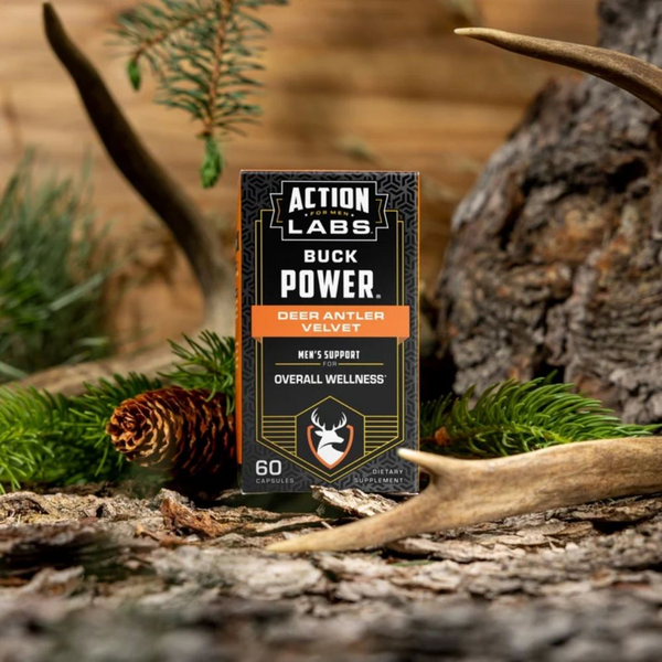 A box of Action Labs Buck Power Deer Antler Velvet dietary supplement stands upright on a forest floor surrounded by pine needles, a pinecone, and antlers.