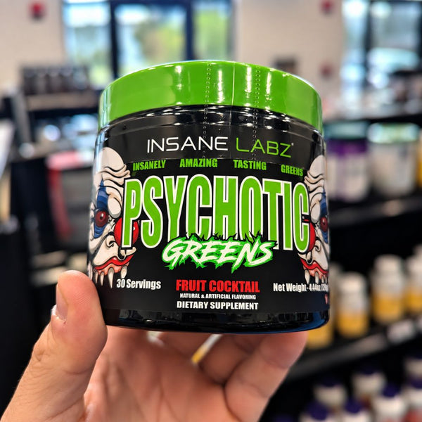 A hand holding a tub of Insane Labz Psychotic Greens dietary supplement in a blurry store setting.